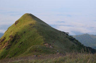 A lush green mountain peak with a narrow path winding up its side. Several hikers are making their way along the trail, surrounded by vast clouds and distant rolling hills.