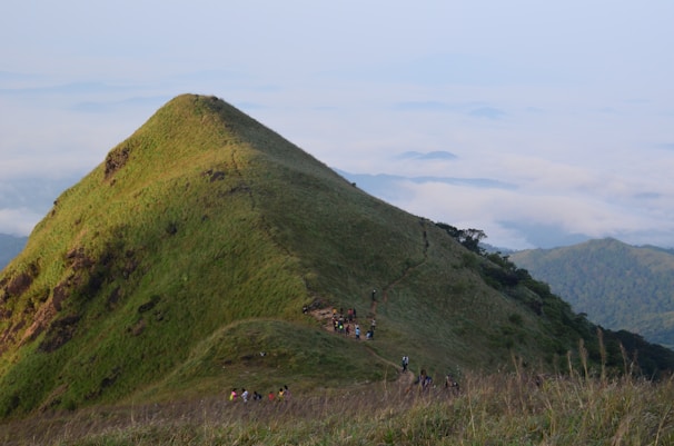 A lush green mountain peak with a narrow path winding up its side. Several hikers are making their way along the trail, surrounded by vast clouds and distant rolling hills.