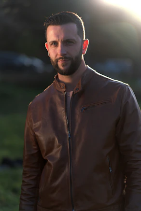 a man wearing a brown leather jacket standing in a field