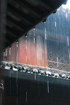 Rain falls heavily, creating visible streaks against a traditional building with intricately designed windows and a tiled roof.