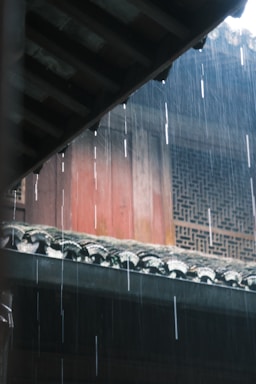 Rain falls heavily, creating visible streaks against a traditional building with intricately designed windows and a tiled roof.