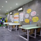 A modern coworking space with several long tables and wooden chairs arranged neatly. The wall features colorful artwork and infographics discussing coworking benefits. Bright colored geometric patterns and text bubbles with statistics are part of the design. White air conditioning units are mounted on the wall, and the room is well-lit with recessed ceiling lights.
