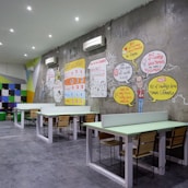 A modern coworking space with several long tables and wooden chairs arranged neatly. The wall features colorful artwork and infographics discussing coworking benefits. Bright colored geometric patterns and text bubbles with statistics are part of the design. White air conditioning units are mounted on the wall, and the room is well-lit with recessed ceiling lights.
