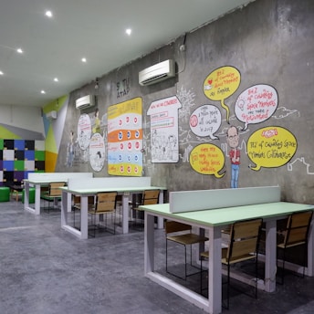 A modern coworking space with several long tables and wooden chairs arranged neatly. The wall features colorful artwork and infographics discussing coworking benefits. Bright colored geometric patterns and text bubbles with statistics are part of the design. White air conditioning units are mounted on the wall, and the room is well-lit with recessed ceiling lights.