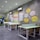 A modern coworking space with several long tables and wooden chairs arranged neatly. The wall features colorful artwork and infographics discussing coworking benefits. Bright colored geometric patterns and text bubbles with statistics are part of the design. White air conditioning units are mounted on the wall, and the room is well-lit with recessed ceiling lights.