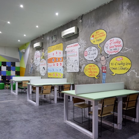A modern coworking space with several long tables and wooden chairs arranged neatly. The wall features colorful artwork and infographics discussing coworking benefits. Bright colored geometric patterns and text bubbles with statistics are part of the design. White air conditioning units are mounted on the wall, and the room is well-lit with recessed ceiling lights.