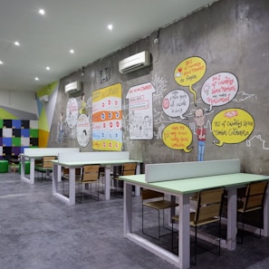 A modern coworking space with several long tables and wooden chairs arranged neatly. The wall features colorful artwork and infographics discussing coworking benefits. Bright colored geometric patterns and text bubbles with statistics are part of the design. White air conditioning units are mounted on the wall, and the room is well-lit with recessed ceiling lights.
