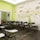 An empty classroom with rows of black chairs featuring attached writing surfaces. The room has a vibrant mural on the wall, depicting a tree with colorful elements and words related to business and creativity. The overall setting is bright with a combination of lime green and white colors on the walls.