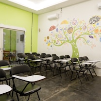 An empty classroom with rows of black chairs featuring attached writing surfaces. The room has a vibrant mural on the wall, depicting a tree with colorful elements and words related to business and creativity. The overall setting is bright with a combination of lime green and white colors on the walls.