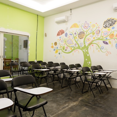 An empty classroom with rows of black chairs featuring attached writing surfaces. The room has a vibrant mural on the wall, depicting a tree with colorful elements and words related to business and creativity. The overall setting is bright with a combination of lime green and white colors on the walls.