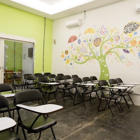 An empty classroom with rows of black chairs featuring attached writing surfaces. The room has a vibrant mural on the wall, depicting a tree with colorful elements and words related to business and creativity. The overall setting is bright with a combination of lime green and white colors on the walls.
