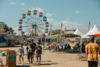 A lively amusement park scene featuring a large Ferris wheel in the background with colorful seats and several other amusement rides. People of various ages can be seen walking and engaging in activities. The setting has tents, stalls with colorful signage, and various other attractions typical of a fairground. The sky is clear with a few scattered clouds.