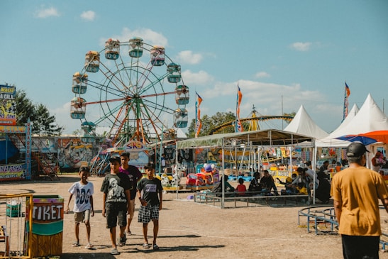 A lively amusement park scene featuring a large Ferris wheel in the background with colorful seats and several other amusement rides. People of various ages can be seen walking and engaging in activities. The setting has tents, stalls with colorful signage, and various other attractions typical of a fairground. The sky is clear with a few scattered clouds.