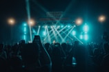 A concert scene with a large crowd of people holding up their phones, capturing the performance. Bright stage lights illuminate the area, with beams of light directed towards the audience. The stage is set up with musical equipment and performers under a large sign lit in blue hues.