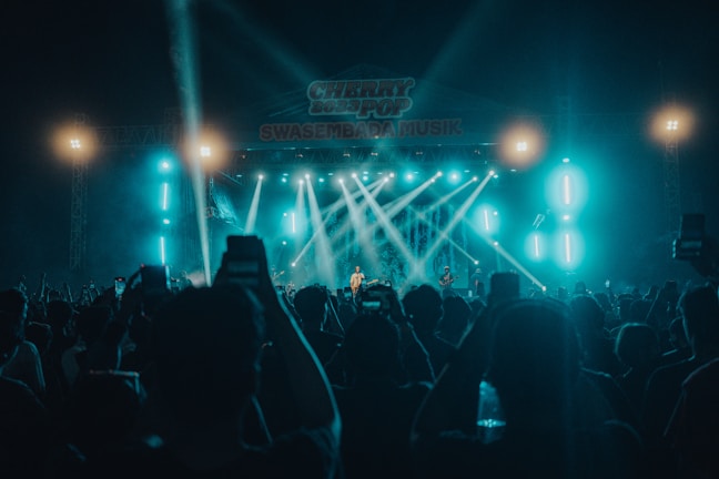 A concert scene with a large crowd of people holding up their phones, capturing the performance. Bright stage lights illuminate the area, with beams of light directed towards the audience. The stage is set up with musical equipment and performers under a large sign lit in blue hues.
