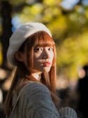 A young woman with long hair is wearing a cozy knitted sweater and a fluffy white beret. The background features a blur of greenery with the sunlight creating a warm glow, highlighting the subject's face and hair.