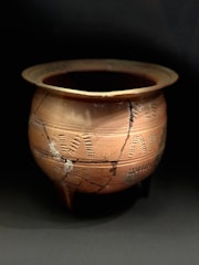 A large, round, terracotta pot with decorative imprints and visible cracks. The pot exhibits signs of aging and repair, suggesting historical or archaeological significance. The lighting highlights the pot's earthy tones and intricate patterns.