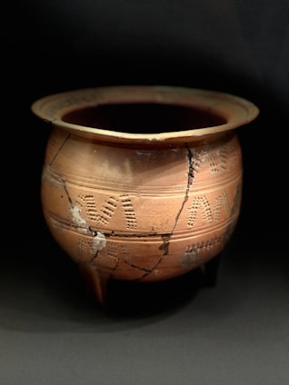 A large, round, terracotta pot with decorative imprints and visible cracks. The pot exhibits signs of aging and repair, suggesting historical or archaeological significance. The lighting highlights the pot's earthy tones and intricate patterns.