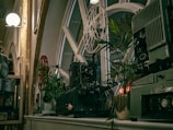 Antique film projectors are placed on a shelf near large window panes with a geometric design. Surrounding the projectors, there are potted green plants adding a natural element. The scene is well-lit with a soft glow from overhead lights enhancing the vintage aesthetic.