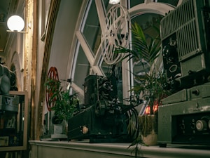 Antique film projectors are placed on a shelf near large window panes with a geometric design. Surrounding the projectors, there are potted green plants adding a natural element. The scene is well-lit with a soft glow from overhead lights enhancing the vintage aesthetic.