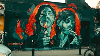 Vibrant street art depicting two female figures on a brick wall, each holding a lighter and cigarette, with a mix of red, blue, and black hues surrounding them. The mural includes intricate details, such as facial expressions and jewelry, and is set against an urban backdrop.