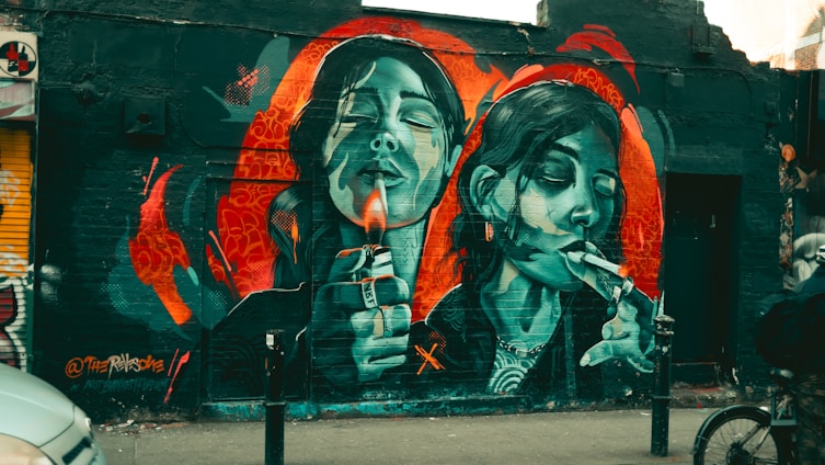 Vibrant street art depicting two female figures on a brick wall, each holding a lighter and cigarette, with a mix of red, blue, and black hues surrounding them. The mural includes intricate details, such as facial expressions and jewelry, and is set against an urban backdrop.