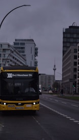 A yellow city bus is positioned in the foreground on a street that runs through a modern urban area. The bus displays the route number 300 and the destination 'S+U Warschauer Str.' A prominent television tower is visible in the background, indicating a metropolitan location. The surrounding buildings are tall and modern, with cloudy skies overhead creating a gloomy atmosphere.