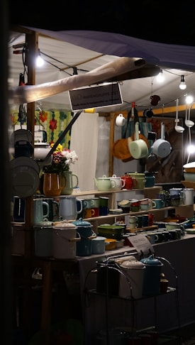A market stall displaying a variety of colorful enamel kitchenware, including teapots, mugs, and pots. Some overhead lights illuminate the area, creating a warm and inviting atmosphere. A small bouquet of flowers adds a touch of freshness to the display.