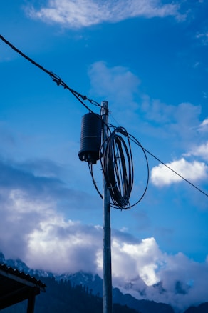 A utility pole with a coiled cable and a cylindrical transformer is set against a backdrop of a vibrant blue sky with scattered clouds. A mountainous landscape with snow-capped peaks is visible in the distance.