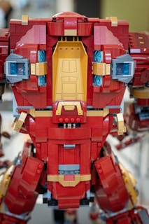 A LEGO model features a large robot or mech suit constructed from red, gold, and grey bricks. The model is detailed with sections that resemble armor plating and mechanical components, including blue translucent pieces simulating lights.