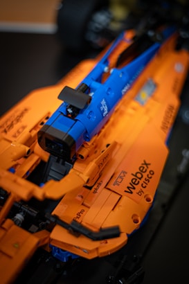 A detailed close-up of a model racing car predominantly colored in orange with parts in blue and black. The car has several visible sponsors' logos, including 'Webex by Cisco' and 'Nissan.' The model appears to be made of interlocking pieces, possibly Lego or a similar construction toy.