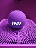 A spherical smart speaker with an LED display showing the time 22:22 is placed on a circular device with a grid-like pattern. The lighting has a pinkish-purple hue, creating a modern and moody atmosphere.