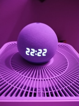 A spherical smart speaker with an LED display showing the time 22:22 is placed on a circular device with a grid-like pattern. The lighting has a pinkish-purple hue, creating a modern and moody atmosphere.