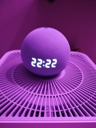 A spherical smart speaker with an LED display showing the time 22:22 is placed on a circular device with a grid-like pattern. The lighting has a pinkish-purple hue, creating a modern and moody atmosphere.