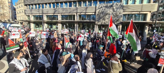 A group of people are gathered for a demonstration outside a modern building. They are holding various signs and flags, including Palestinian flags and signs with slogans supporting Gaza and Palestine. The sun casts shadows on the building and people, and the street is filled with activity.