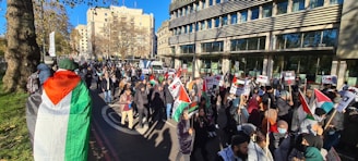 A large group of people are gathered for a protest, holding Palestinian flags and signs with messages related to Gaza and Palestine. The crowd is moving along a city street lined with buildings, and it appears to be a sunny day.