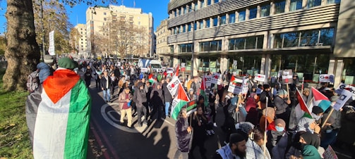 A large group of people are gathered for a protest, holding Palestinian flags and signs with messages related to Gaza and Palestine. The crowd is moving along a city street lined with buildings, and it appears to be a sunny day.