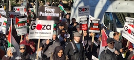 A large group of people are participating in a protest, holding various signs with messages related to Palestine, including 'Free Palestine,' 'Stop the Massacre,' and 'Stop Gaza Genocide.' Many are wearing scarves with black and white patterns. The gathering appears to be peaceful with individuals focused on the cause they are supporting.