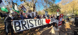 A group of people are gathered outdoors holding flags and signs, including a large banner reading 'BRISTOL ❤️'. The scene is set in a park-like area with autumn leaves scattered on the ground and trees around. The crowd appears to be participating in a demonstration or march. Various flags and signs indicate a focus on political or social issues.