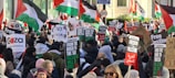 A large crowd of people is gathered, holding signs and flags with messages supporting Palestine. The scene is vibrant with many individuals wearing winter clothing, indicating a protest or demonstration. Palestinian flags dominate the background, and various posters convey messages such as 'Free Palestine' and other pro-Palestinian sentiments.