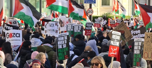 A large crowd of people is gathered, holding signs and flags with messages supporting Palestine. The scene is vibrant with many individuals wearing winter clothing, indicating a protest or demonstration. Palestinian flags dominate the background, and various posters convey messages such as 'Free Palestine' and other pro-Palestinian sentiments.
