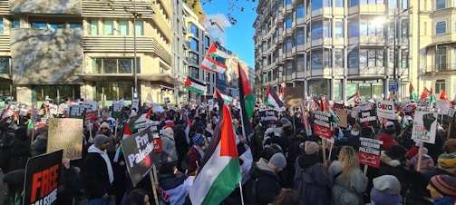 A large crowd gathered in an urban setting, holding flags and protest signs advocating for Palestinian freedom. The atmosphere is charged with activism as people stand closely together, filling the street against a backdrop of modern buildings. Many of the signs prominently display messages of support for Palestine.