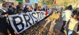 A group of people are holding a large banner with the word 'BRISTOL' written on it, participating in what appears to be a public demonstration or protest. The crowd includes people of various ages, dressed for cool weather, and some are holding signs. Flags are visible, and the setting is outdoors on a pathway covered with autumn leaves, with trees in the background.