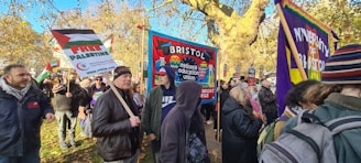 A group of people are gathered outdoors, holding various banners and signs. The surroundings include trees with autumn leaves and a clear blue sky. One prominent sign reads 'FREE PALESTINE.' Another banner features the words 'Bristol National Education Union.' People are dressed warmly, suggesting cooler weather.