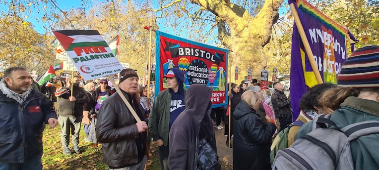 A group of people are gathered outdoors, holding various banners and signs. The surroundings include trees with autumn leaves and a clear blue sky. One prominent sign reads 'FREE PALESTINE.' Another banner features the words 'Bristol National Education Union.' People are dressed warmly, suggesting cooler weather.