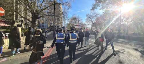 A group of people walking along a city street, some carrying signs with messages. The scene includes police officers in uniform, wearing reflective vests, positioned near the crowd. Leafless trees and buildings are visible in the background, with sunlight streaming through the branches, creating a bright glare.