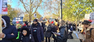 A group of people is gathered outdoors, holding signs and banners with messages related to political activism. The scene takes place on a sunny day with trees in the background showing autumn colors. Many people are wearing jackets, indicating cool weather.
