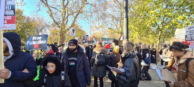 A group of people is gathered outdoors, holding signs and banners with messages related to political activism. The scene takes place on a sunny day with trees in the background showing autumn colors. Many people are wearing jackets, indicating cool weather.