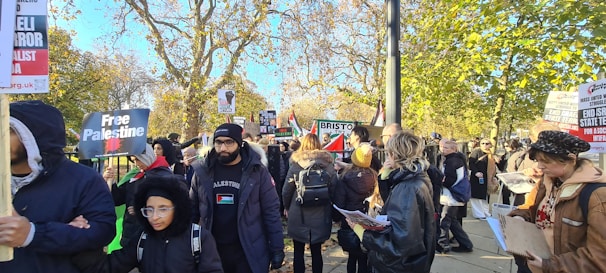 A group of people is gathered outdoors, holding signs and banners with messages related to political activism. The scene takes place on a sunny day with trees in the background showing autumn colors. Many people are wearing jackets, indicating cool weather.