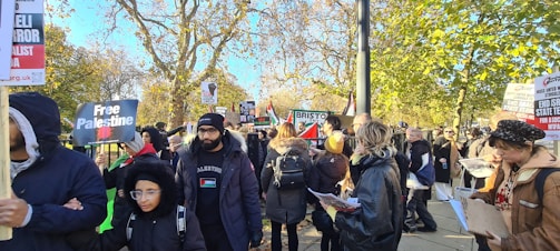 A group of people is gathered outdoors, holding signs and banners with messages related to political activism. The scene takes place on a sunny day with trees in the background showing autumn colors. Many people are wearing jackets, indicating cool weather.